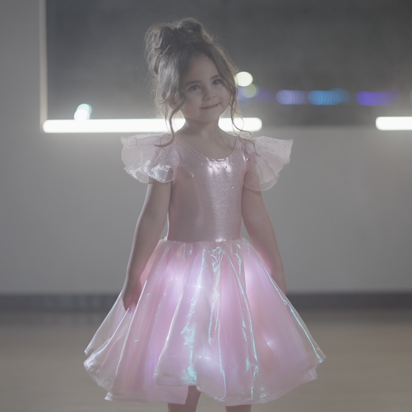 A young girl stands in a dimly lit room wearing a sparkly pink tutu dress with shimmering details.