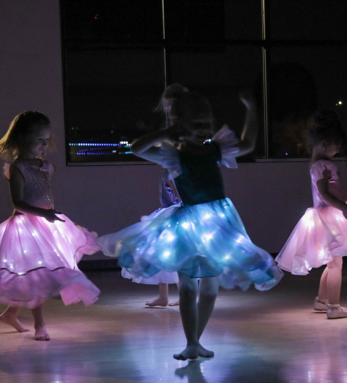Three young girls dancing in illuminated tutus inside a dark room. The center girl wears a glowing blue tutu, while the girls on the left and right wear glowing pink tutus.